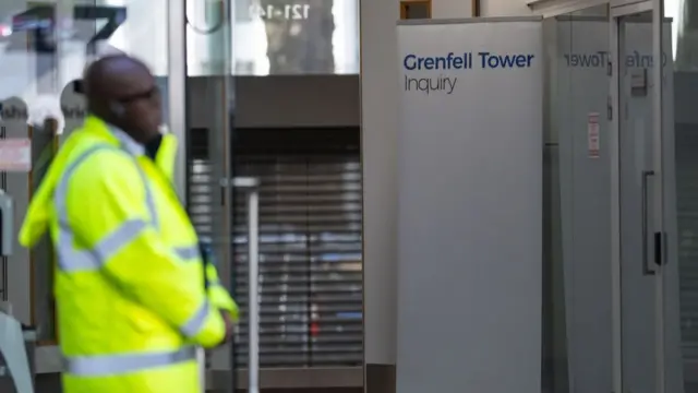 A security officer in hi-vis stands at the entrance to Dorland House, a sign in the background reads 'Grenfell Tower Inquiry'