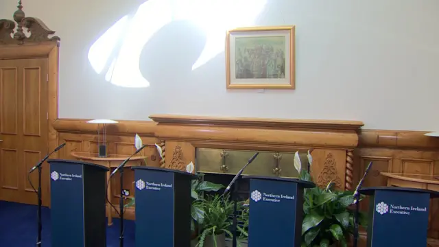 Blue podiums with 'Northern Ireland Executive' in white font. The four podiums are placed in front of a light wooden fireplace.