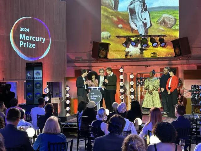 English Teacher, a four-person group, accepts the Mercury Prize on stage with people sitting at tables in the forefront