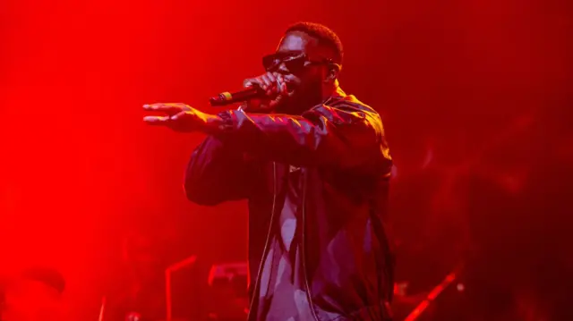 Ghetts is wearing black square sunglasses and a black jacket while singing into a microphone with his hand outstraged. There is red lighting on him.