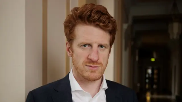 Matthew O'Toole - a man with red hair and a beard looks at the camera while wearing a dark coloured blazer and white collared shirt. He is standing in a hallway with white walls.