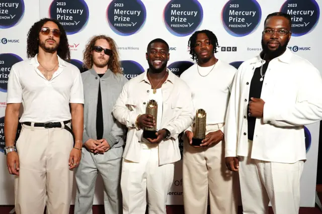 the five members of Ezra Collective pose for a photo, all wearing white and beige tops and trousers, except one member who is wearing grey. Two of them are holding an award.