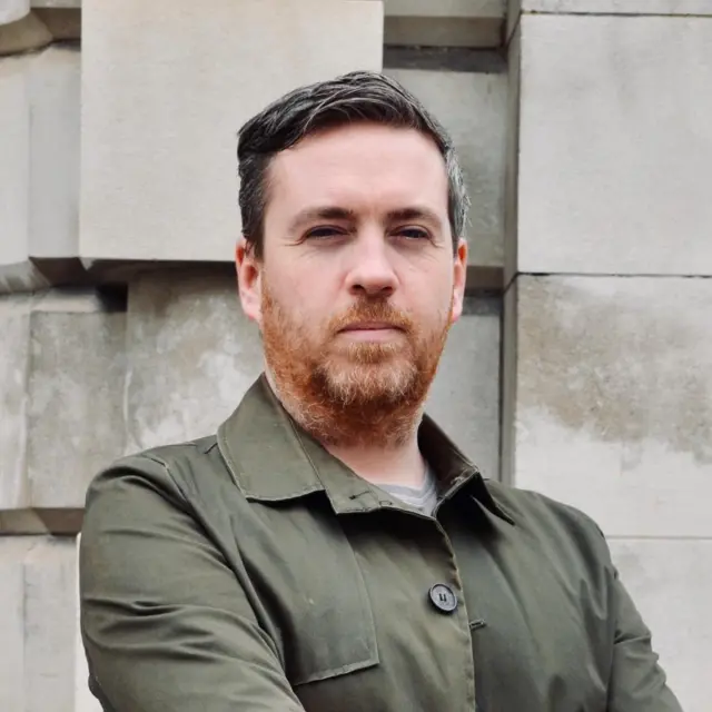 A man with black hair and a ginger beard looks into directly into the camera, his mouth his closed. He is wearing a green jacket and his arms are folded