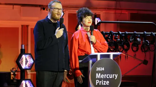 Huw Stephens and Annie Mac hold microphones on the stage in front of a podium that has written on it 2024 Mercury Prize