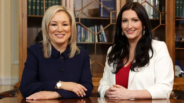 Michelle O'Neill and Emma Little-Pengelly - two women, one with mid-length blonde hair wearing a navy button coat and another with dark, longer hair wearing a white blazer and red top - smile at the camera while sitting down. They are in front of a glass bookcase.