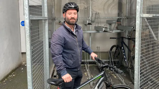 A man stands holding a black bicycle outside of a box with two bicycles in it. The man is wearing black jeans, a navy bomber jacket, and a black helmet. He is looking at the camera.