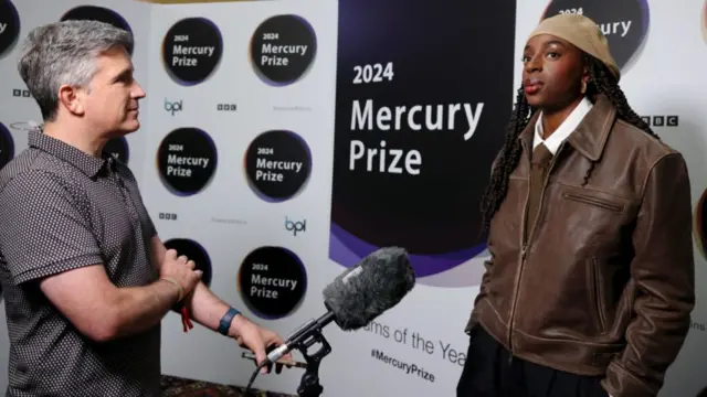 Cat Burns stands in front of a board with the words 2024 Mercury Prize written on it. An interviewer stands in front of her holding a microphone