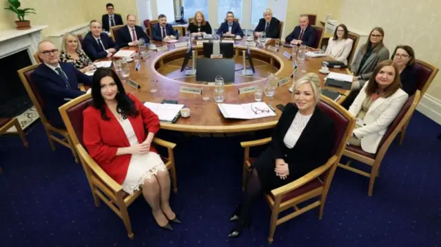 A number of politicians sit around a circular table in the first NI Executive meeting in February 2024. The room has blue carpet and light print wallpaper.