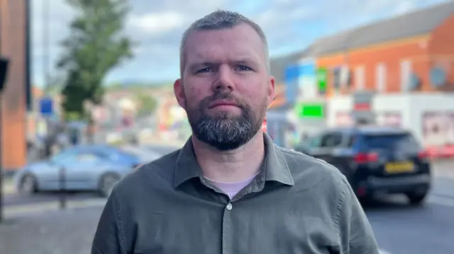 Gerry Carroll - a man with short hair and a beard stands in the middle of a footpath looking at the camera. He is wearing a green button up shirt and a light coloured t-shirt..