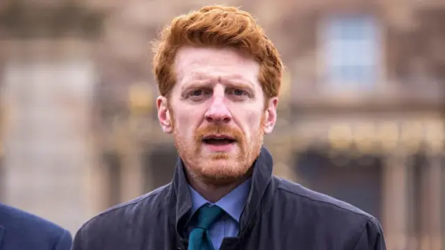 Man with red hair and beard looks directly into the camera. He is wearing a green tie and dark jacket