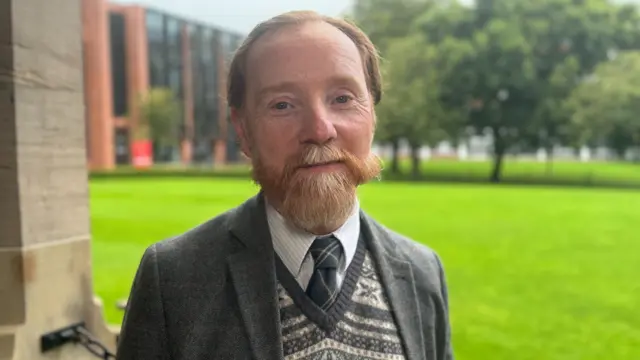 A man with a red-orange beard and flowing moustache wearing a patterned, knitted jumper and dark grey jacket looks at the camera.