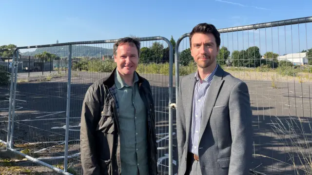 Two men standing in front of an empty site, one is wearing a suit with black hair and the other is wearing a shirt and jacket he also has brown hair