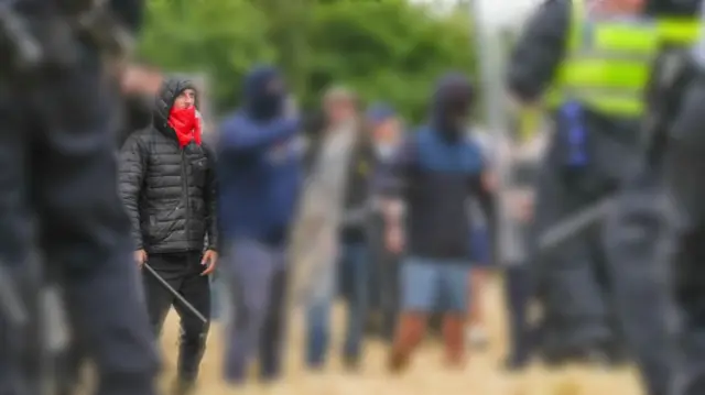 Thomas Birley holding a baton with a mask covering half his face, with others around him blurred out