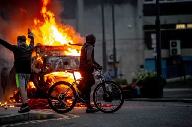 A file photo of disorder shows a car burning and two people watching on, one with their face covered