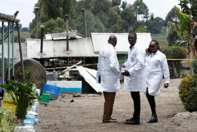 Three people in white coats stand outside the ruined building.