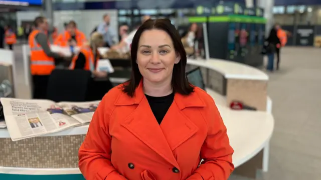 A woman in a dark orange jacket stands in a train station, smiling at the camera. She has shoulder-length dark hair.