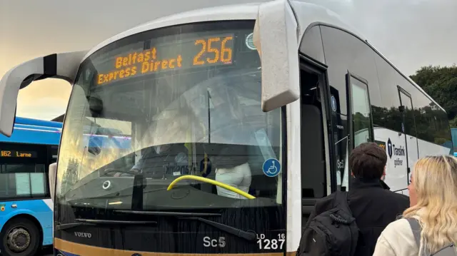 A white Translink bus. Above the windscreen, a display says 'Belfast Express Direct 256'. Two people queue for the bus.