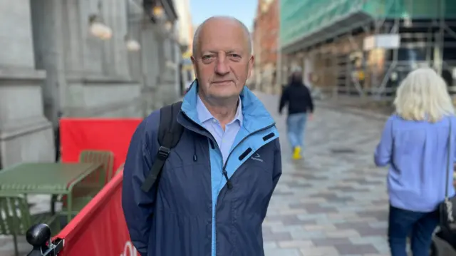 A man stands in a Belfast street, facing the camera. He is wearing a navy jacket.