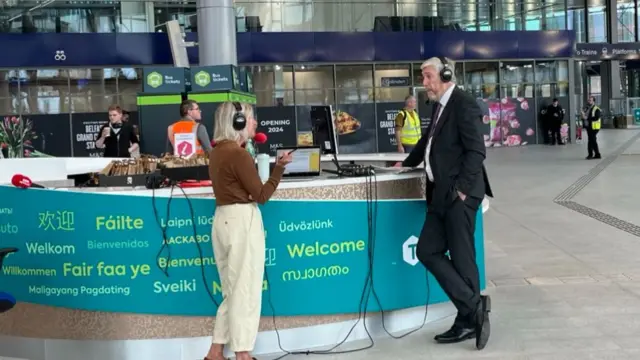 A man and a woman stand facing each other. The woman is dressed in beige trousers and a brown shirt. The man is dressed in a black suit with a white shirt. They are standing in a train station.