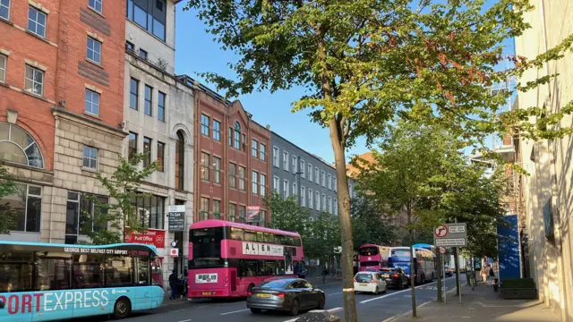 A street in Belfast. On the far side of the street, there are three buses. The first bus is light blue, while the other buses are pink double-deckers.