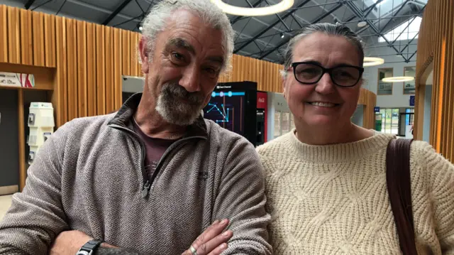 A man wearing a grey jumper and woman wearing a cream jumper, she has glasses