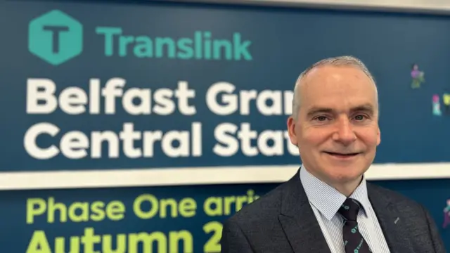 A man stands smiling at the camera in front of a sign which reads 'Translink - Belfast Grand Central Station'. The man is wearing a dark suit with a tie with numerous Translink logos.