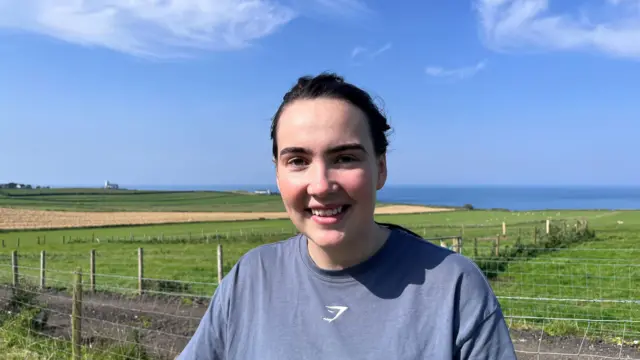 A woman smiling at the camera. She is wearing a dark blue t-shirt. There are fields of grass and a clear blue sky in the background.