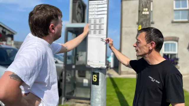 Two men standing looking at a bus timetable on a lampost. The man on the left has a mullet and is wearing a white t-shirt. The man on the right has short black hair and is wearing a black t-shirt.