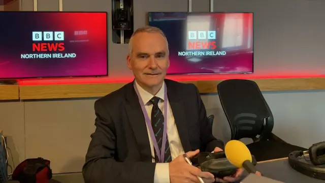 Chris Conway in a suit sits in a radio studio, looking at the camera.