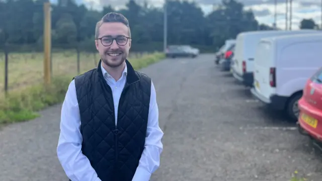 A man wearing a shirt and gilet, he has glasses and brown hair