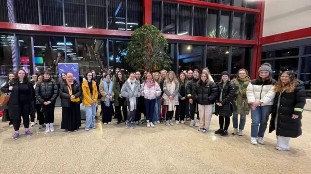 A group of about thirty people stand in a large foyer. It is pitch black outside, and all of the people are dressed in cold-weather clothing.