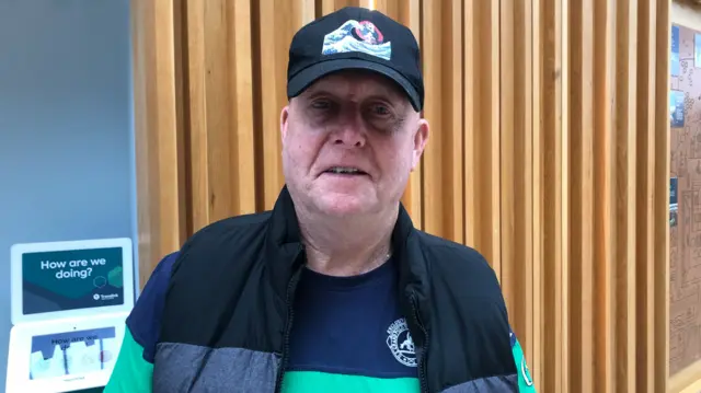 A man wearing a black cap and a black gilet, he has a green and navy t shirt on