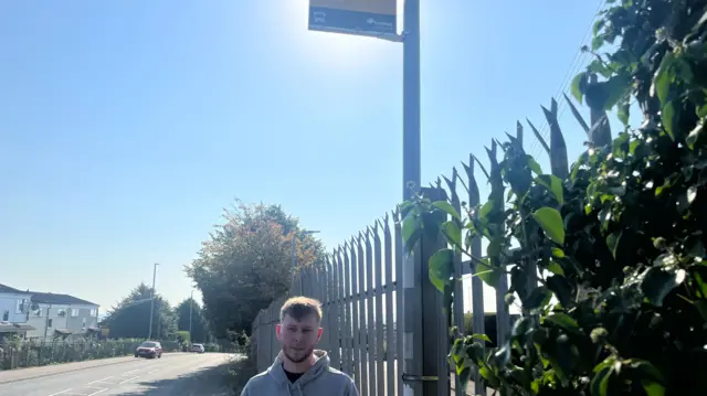 A man standing at a bus stop, he has dark hair and a dark beard