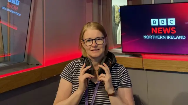A woman in a black and white t-shirt looks at the camera. She is sat in a studio in front of a screen displaying the 'BBC NEWS NORTHERN IRELAND' logo