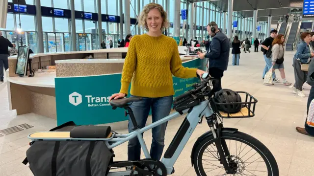 A woman stands inside Belfast's Grand Central Station with a blue electric bike in front of her. She has blonde hair and is smiling at the camera.