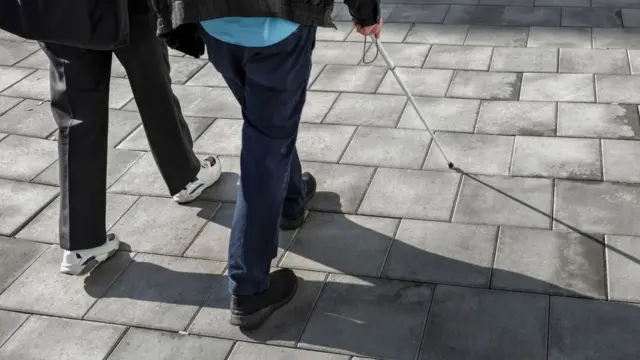 The picture shows two sets of legs on grey paving stones, with one person using a white mobility cane.