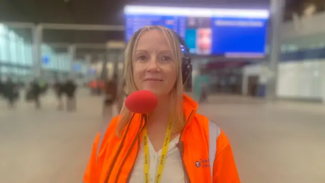 A woman with blonde hair dressed in a white shirt and a high-vis orange jacket smiles at the camera. She is wearing a headset.