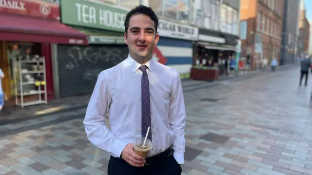A man stands on a street, facing the camera. He is wearing a white shirt and holding a drink in a clear plastic container.