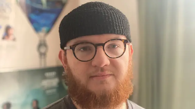 A close-up of a man with an auburn beard. He is wearing glasses and a black beanie.