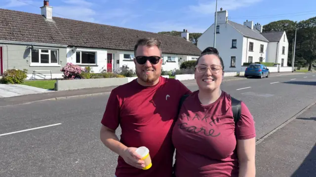 A man and a woman standing on the side of a road. They are both smiling and both have red t-shirts on.