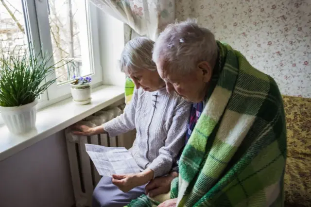 A senior woman holding gas bill in front of heating radiator as elderly man sits next to her wrapped in a green blanket.. Payment for heating in winter.