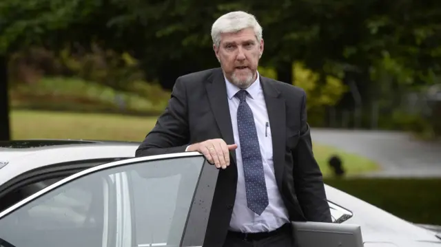 A man stands beside a car. He is wearing a dark suit, with a navy tie. He has short white hair.