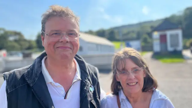 A man and a woman looking at the camera and smiling. Both are wearing white tops and glasses.