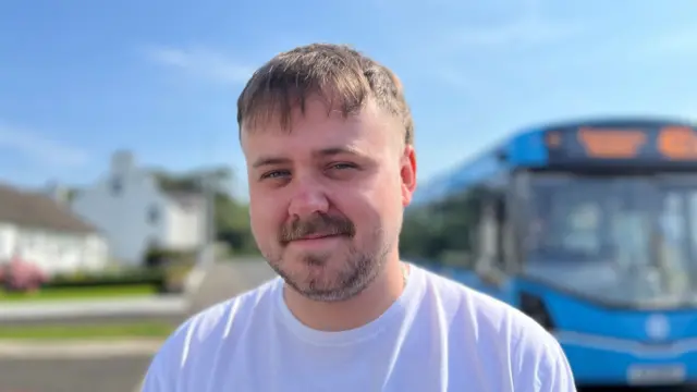 A man stands smiling at the camera. He has a mullet, and is wearing a white t-shirt. There is a bus behind him.