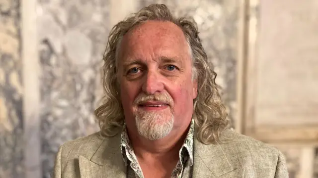 Michael Stewart wearing a cream suit, he has a white beard and blonde hair