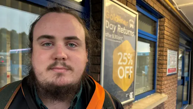 A man stands, looking at the camera. Behind him is an advert for a bus ticket.