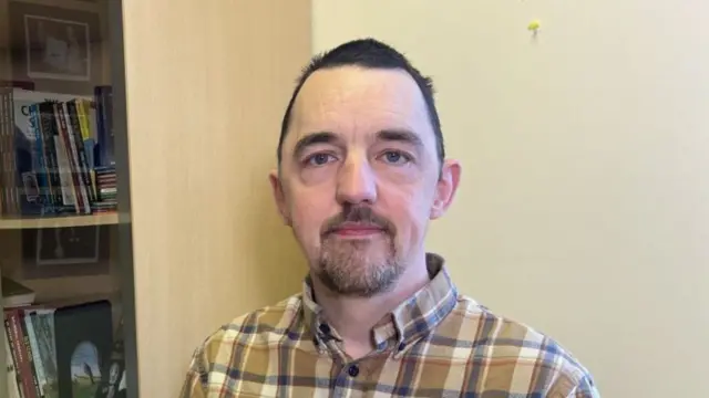 A man in a brown, grey, and black shirt looks at the camera. He has short black hair.