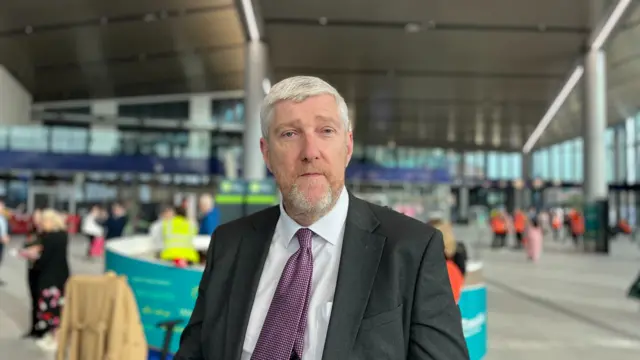 A man stands in a train station, facing the camera. He is wearing a black suit with a white shirt and a purple tie. He has short white hair.