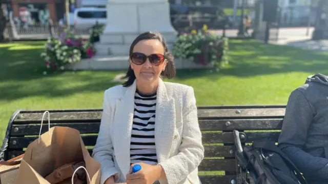 A woman sits on a bench, facing the camera. She is wearing a cream jacket and a black and white shirt. She is also wearing sunglasses, and has dark hair.