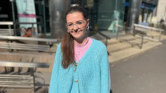 A woman stands in a Belfast street, facing the camera. She is wearing a pink jumper and a turquoise jumper. She has brown hair and glasses.